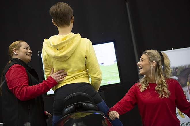 Kuva Liikuntamaasta 2019. 2 punapaitaista iloista naista opastaa nuorta ratsastussimulaattorilla