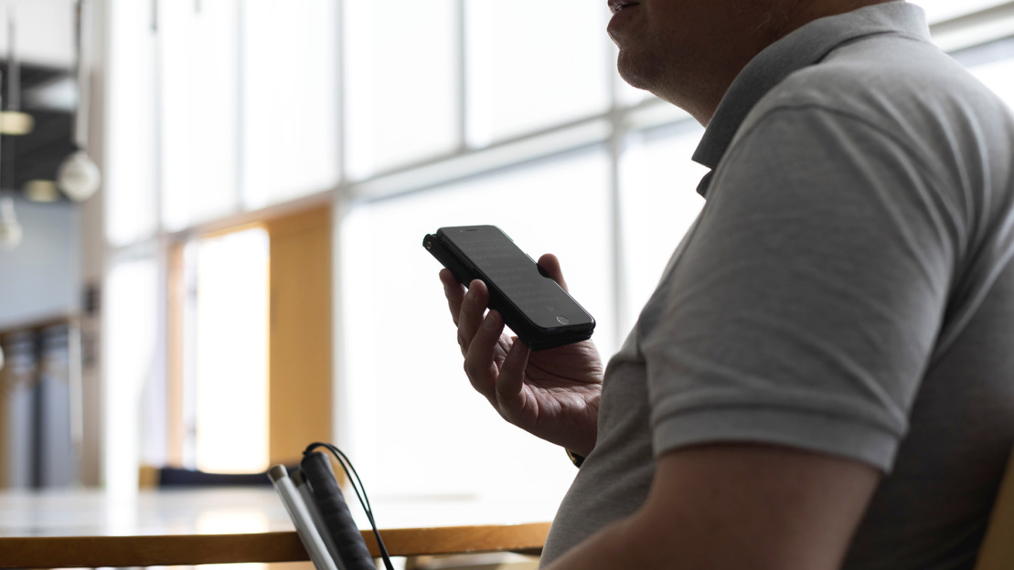 Kuvassa näkövammaien mies istuu tuolilla, valkoinen keppi toisessa kädessä, älypuhelin toisessa. Puhelin on kohotettuna, hän kuuntelee sieltä jotain.