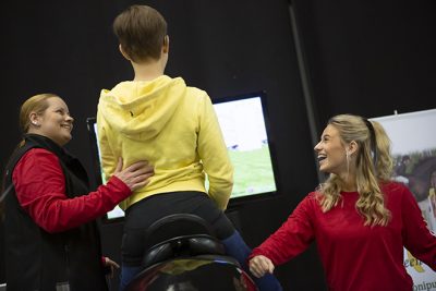 Kuva Liikuntamaasta 2019. 2 punapaitaista iloista naista opastaa nuorta ratsastussimulaattorilla