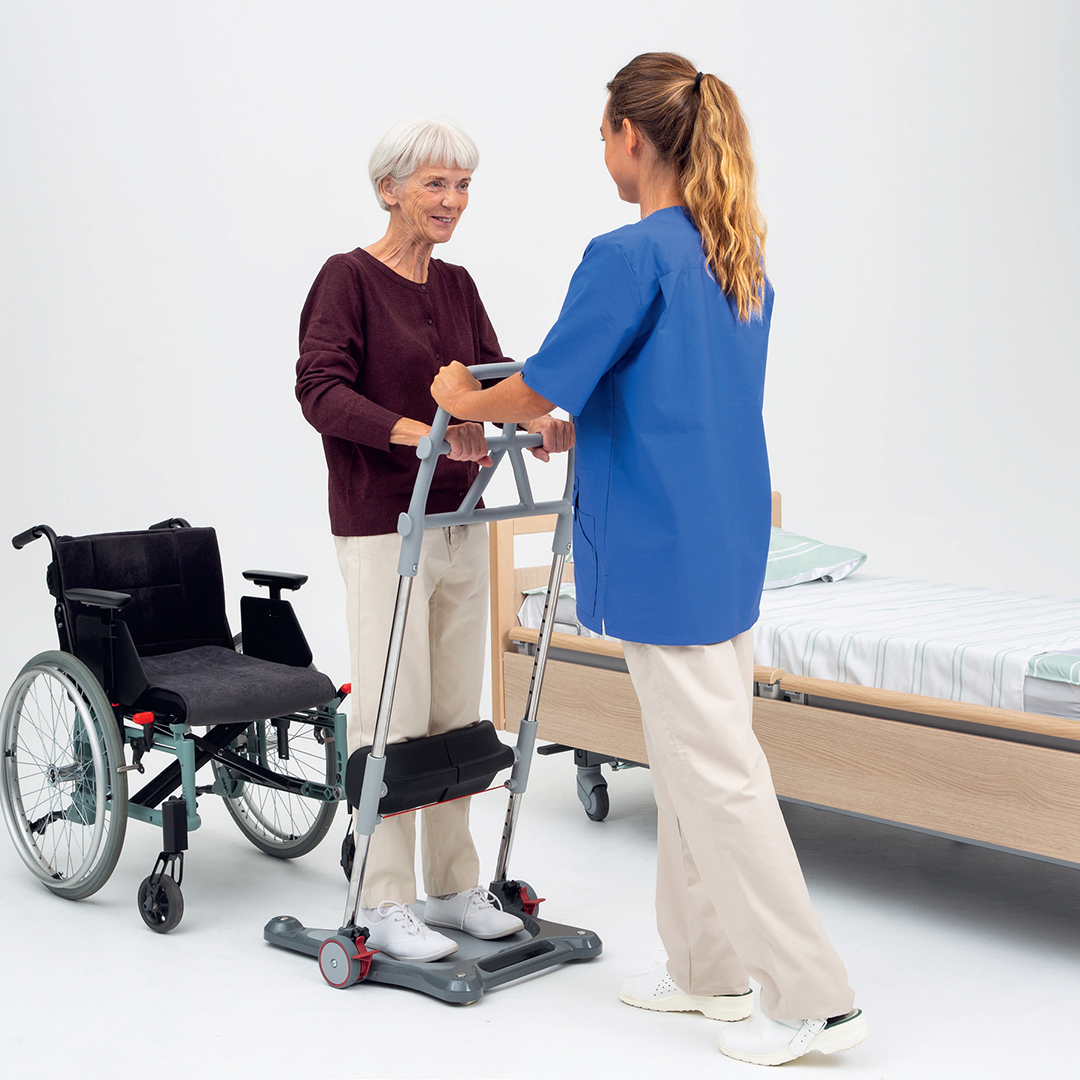 Kuvassa hoitaja avustaa ikääntynyttä siirtymään pyörätuolista sänkyyn siirtotelineen avulla. Kuva: Berner Medical, Molift Raiser Pro -siirtoteline