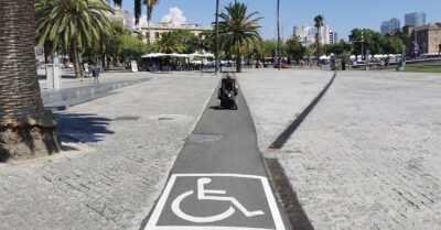 Kuva etelän auringosta, matkaaja kulkee pyörätuolilla torin poikki, missä mukulakivien keskelle on tehty asvaltoitu väylä, mihin maalattu pyörätuoli-merkki.